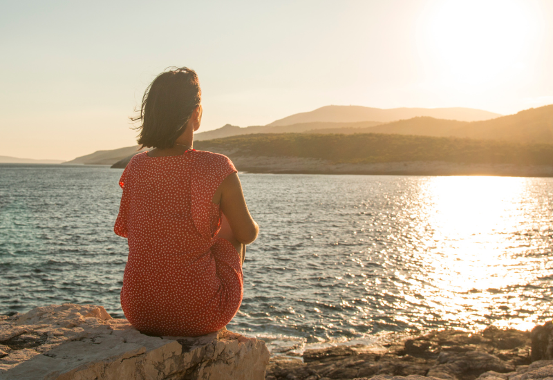 Person sitting by the water at sunset, reflecting quietly and practicing being kind to yourself.